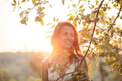 Smiling mature woman standing by tree against sky during sunset