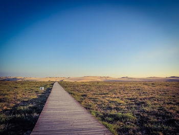 Boardwalk leading towards field against clear blue sky