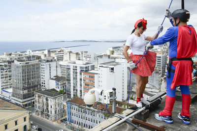 Woman wearing hero costume being prepared by a man to descend a tall rappel building. 