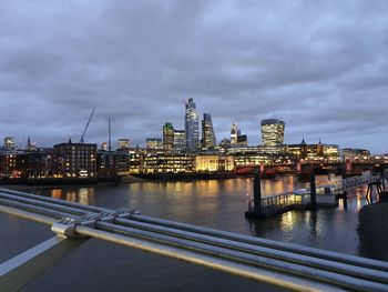 Illuminated buildings by river against cloudy sky