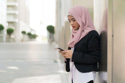 Businesswoman wearing hijab using mobile phone while standing against building