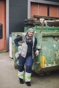 Full length portrait of smiling female construction worker standing with hand on hip by metal container
