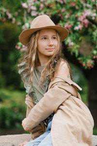 Caucasian teenage girl with long blonde hair in a trench coat sitting on a rock in the park