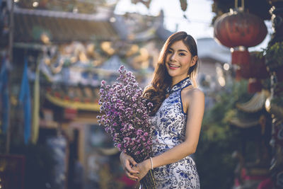 Portrait of smiling young woman holding flowers while standing outdoors