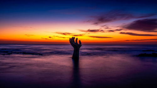 Cropped hand emerging from sea against dramatic sky during sunset