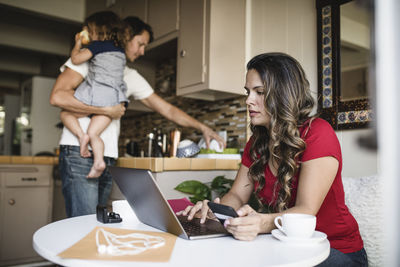 Mother using laptop while father carrying daughter and working in kitchen