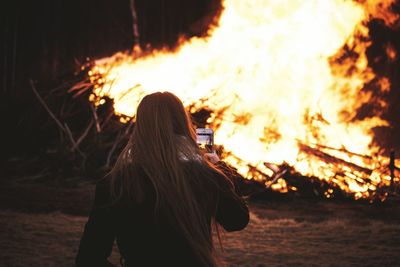 Rear view of woman photographing bonfire with mobile phone at night