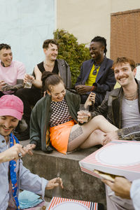 Happy non-binary friends celebrating with food and drinks while sitting on bench