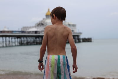 Rear view of shirtless boy standing at beach against sky