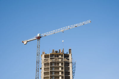 Low angle view of crane by building against clear blue sky