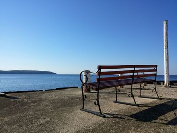 Empty bench by sea against clear blue sky