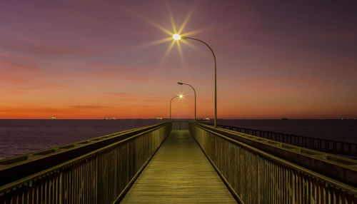 Illuminated street light on pier amidst sea against sky during sunset
