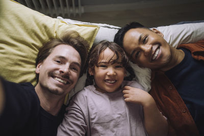 Directly above portrait of happy gay couple lying down with daughter at home