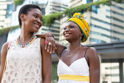Cheerful adult african american female best friends in stylish summer dresses and authentic accessories laughing while walking on city street near contemporary skyscrapers