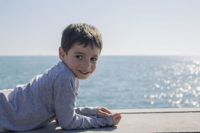 Smiling boy looking away while lying on retaining wall against sea and sky