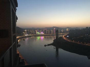 Illuminated bridge over river by city buildings against sky during sunset