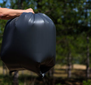 Cropped hand of man holding black garbage bag against trees