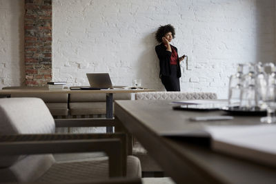 Confident young female lawyer talking on mobile phone while leaning on white wall at office