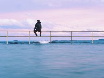 Rear view of man sitting on railing amidst sea