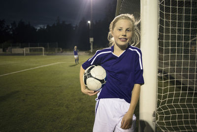 Portrait of happy girl holding soccer ball standing by goal post on field