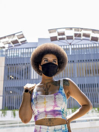 Confident african american female in medical mask and fashionable summer outfit standing in urban area in city during coronavirus outbreak