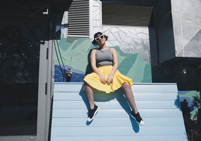 Low angle portrait of young woman sitting on bench against mosaic wall