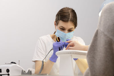 White nail technician in mask drills, removes client's cuticle with hardware machine, holding nail