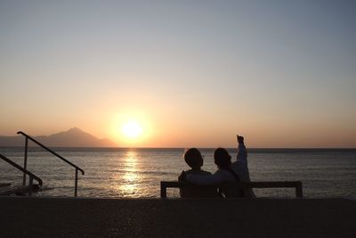 Men sitting on shore against sky during sunset