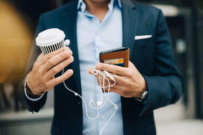 Midsection of businessman holding smart phone with disposable coffee cup and in-ear headphones while standing in city