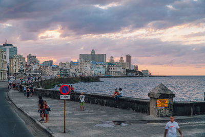 People on city buildings by sea against sky during sunset