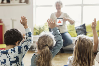 Rear view of students raising hands while teacher showing digital tablet at preschool