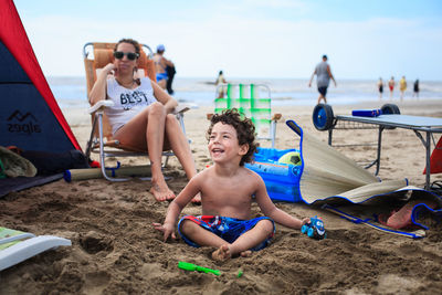 Mother looking at shirtless son sitting with toys on beach