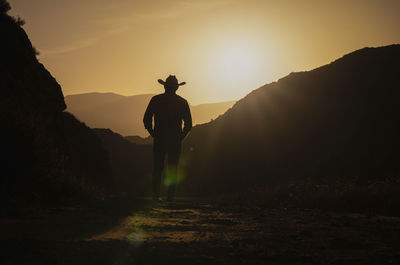 Silhouette of adult man in cowboy hat against mountain and sky during sunset