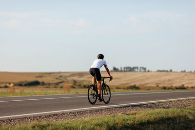 Back view of professional cyclist on white t-short and black shorts riding bike on paved road. 