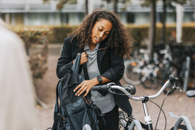 Businesswoman talking on smart phone holding backpack while standing next to bicycle