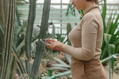 A beautiful plus size girl uses smartphone standing among the green plants of the greenhouse.