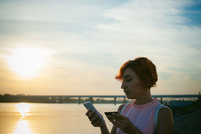 Mid adult woman using mobile phone while standing by river against sky during sunset