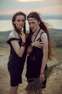 Two woman in black dresses walk on the mountain with a bouquet of flowers in summer at sunset