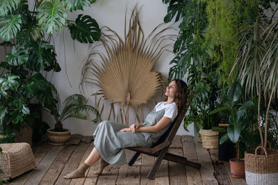 Young woman relaxed sit on comfortable armchair in stylish indoor garden with green exotic plants