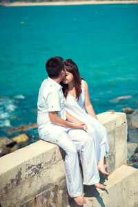 Man kissing woman on forehead while sitting on retaining wall against sea