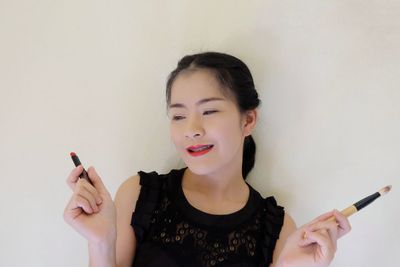 Smiling young woman holding make-up brush and lipstick against wall