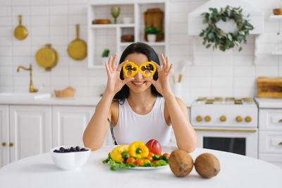 Happy woman nutritionist holds pepper slices to her eyes like glasses, sitting at a table