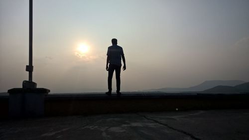 Rear view of man standing on retaining wall against sky during sunset