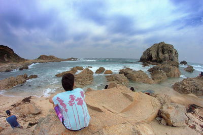 Rear view of woman sitting on rock by sea against sky