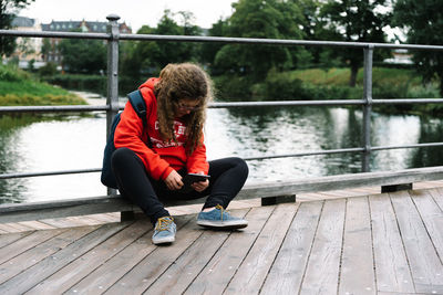 Full length of woman digital tablet while sitting on railing at footbridge over river