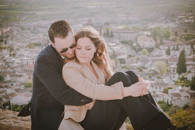 Man embracing woman while sitting on retaining wall against cityscape