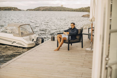 Full length of man reading book while sitting on patio against lake