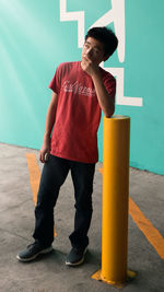 Young man thinking while leaning on bollard while standing on footpath against wall