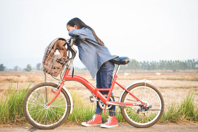 Side view of teenage girl with bicycle on roadside by field against clear sky