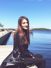 Portrait of smiling teenage girl sitting on pier over river against sky